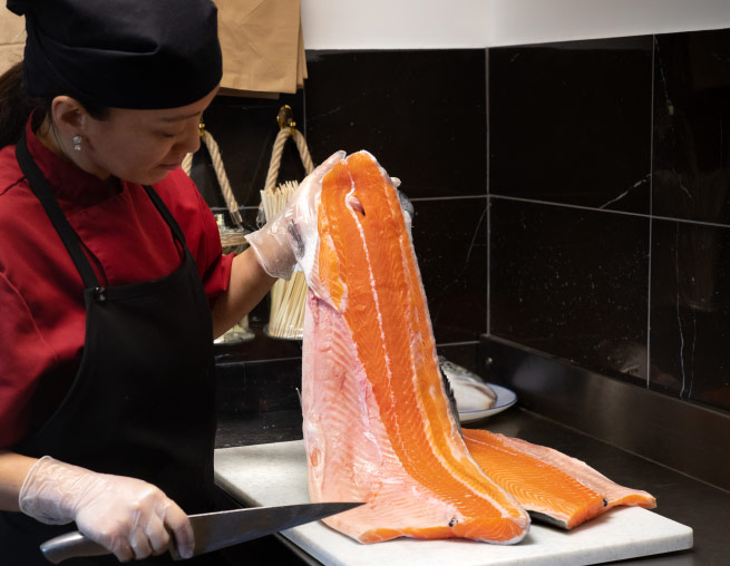 une cuisinière avec un tablier noir tient à la main un immense filet de saumon.